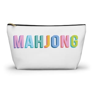 May include: A white zippered pouch featuring the word MAHJONG printed in large, three-dimensional block letters. Each letter displays a different pastel color, including light purple, blue, teal, yellow, and pink, with a black zipper closure.