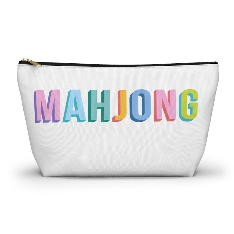 May include: A white zippered pouch featuring the word MAHJONG printed in large, three-dimensional block letters. Each letter displays a different pastel color, including light purple, blue, teal, yellow, and pink, with a black zipper closure.