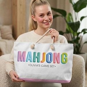 May include: A white canvas tote bag with rope handles featuring the word MAHJONG in large, colorful 3D-style block letters. Below the word, the text reads OLIVIA'S GAME SET in black, with a thin pink horizontal stripe along the bottom edge.