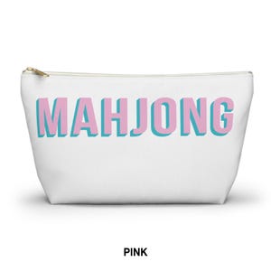 May include: A white zippered pouch featuring the word MAHJONG printed in large, blocky pink letters with a light blue drop shadow effect. The accessory has a gold-toned metal zipper pull at the top left corner and a simple, tapered rectangular shape.