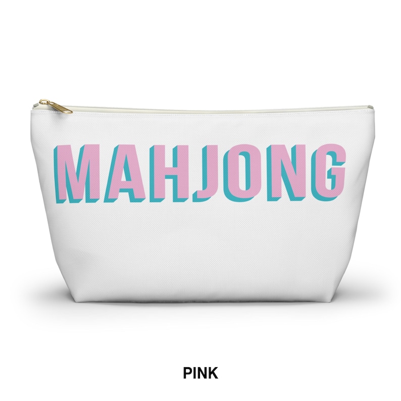 May include: A white zippered pouch featuring the word MAHJONG printed in large, blocky pink letters with a light blue drop shadow effect. The accessory has a gold-toned metal zipper pull at the top left corner and a simple, tapered rectangular shape.