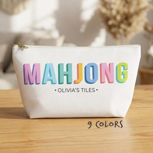 May include: A white zippered pouch featuring the word MAHJONG in large, multi-coloured 3D-style block letters. Below the main text, the words OLIVIA'S TILES are printed in black, flanked by two small dots. The text 9 COLOURS appears at the bottom.