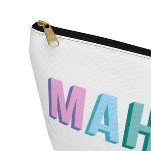 May include: A white zippered pouch featuring the partial text MAH in three-dimensional block letters. The letter M is pink, the letter A is light blue, and the letter H is teal. The pouch has a black zipper track and a gold-toned metal zipper pull.