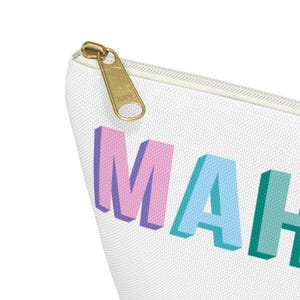 May include: A white zippered pouch featuring the partial text MAH in three-dimensional block letters. The letter M is pink, the letter A is light blue, and the letter H is teal. The pouch has a gold-toned metal zipper pull with a textured finish.