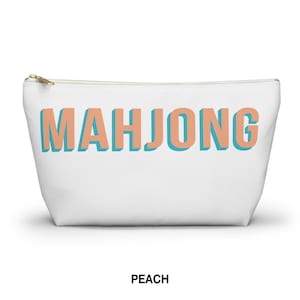 May include: A white zippered pouch featuring the word MAHJONG printed in large, block-style letters. The letters are peach-colored with a teal drop shadow effect. The pouch has a gold-toned metal zipper pull at the top left corner.