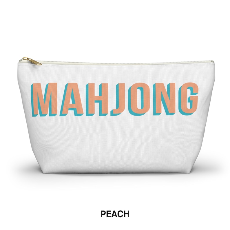 May include: A white zippered pouch featuring the word MAHJONG printed in large, block-style letters. The letters are peach-colored with a teal drop shadow effect. The pouch has a gold-toned metal zipper pull at the top left corner.