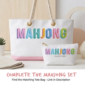 May include: A white canvas tote bag and a matching zippered pouch, both featuring the word MAHJONG in colorful, block-style 3D lettering. The tote has thick rope handles with gold-tone grommets and a pink base. The pouch includes the text OLIVIA'S TILES.