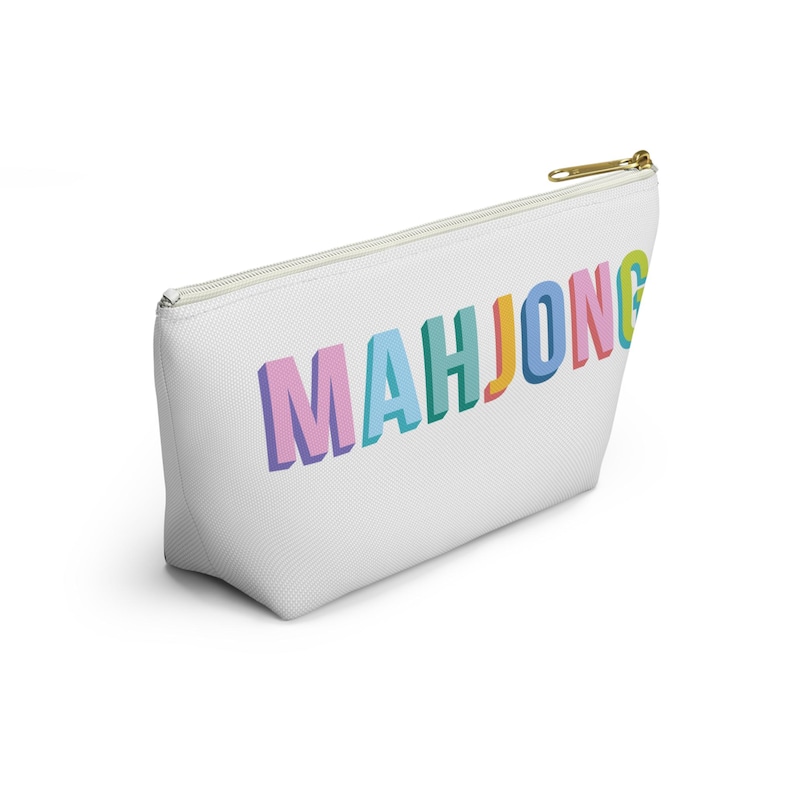 May include: A white zippered pouch featuring the word MAHJONG printed in a three-dimensional, multi-colored block font. The letters are colored in shades of pink, light blue, teal, orange, and yellow, with a gold-toned metal zipper pull.
