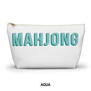May include: A white zippered pouch featuring the word MAHJONG printed in a bold, three-dimensional aqua blue font across the center. The accessory has a gold-toned metal zipper pull at the top left corner and a clean, structured trapezoidal shape.