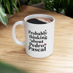 Puede incluir: Taza de cer&aacute;mica blanca con el texto "Probably thinking about Pedro Pascal" en escritura negra. La taza tiene un asa curva y est&aacute; llena de l&iacute;quido oscuro. Un peque&ntilde;o coraz&oacute;n est&aacute; al lado del nombre.
