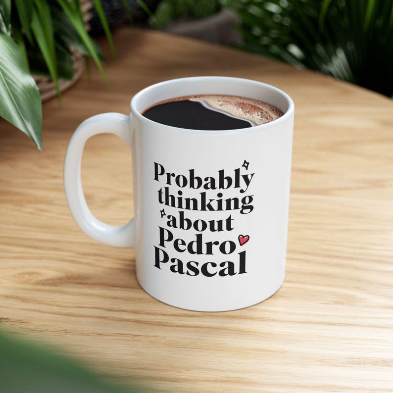 Puede incluir: Taza de cer&aacute;mica blanca con el texto "Probably thinking about Pedro Pascal" en escritura negra. La taza tiene un asa curva y est&aacute; llena de l&iacute;quido oscuro. Un peque&ntilde;o coraz&oacute;n est&aacute; al lado del nombre.