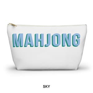 May include: A white zippered pouch featuring the word MAHJONG printed in large, light blue block letters with a dark teal drop shadow effect. The bag has a gold-toned metal zipper pull at the top left corner and a simple, clean rectangular shape.