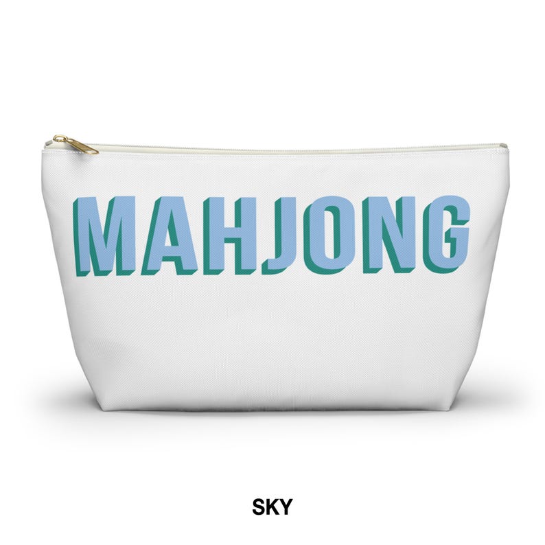 May include: A white zippered pouch featuring the word MAHJONG printed in large, light blue block letters with a dark teal drop shadow effect. The bag has a gold-toned metal zipper pull at the top left corner and a simple, clean rectangular shape.