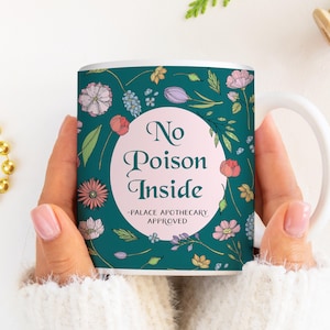 Könnte beinhalten: Weiße Keramiktasse mit einem tealfarbenen Blumendesign und den Worten "No Poison Inside" in einem rosa Kreis. Die Tasse trägt auch den Text "-PALACE APOTHECARY APPROVED".