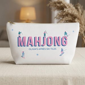 Peut inclure: Une pochette zippée blanche avec le mot MAHJONG écrit en grandes lettres roses à l'ombre bleue. En dessous, le texte indique OLIVIA'S APRÈS SKI TILES. Le motif comprend de petites illustrations de skieurs et des flocons bleu clair.