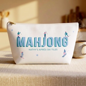 May include: A white zippered pouch featuring the blue text MAHJONG and KATHY'S APRÈS SKI TILES. The design includes small illustrations of people skiing in colourful winter gear and light blue snowflake accents scattered across the fabric surface.