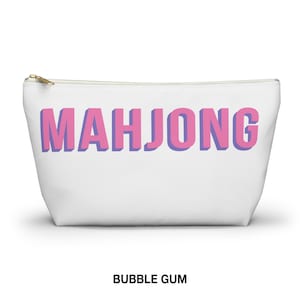 May include: A white zippered pouch featuring the word MAHJONG printed in large, blocky pink letters with a purple shadow effect. The accessory has a gold-toned metal zipper pull at the top left corner and a clean, minimalist design throughout.