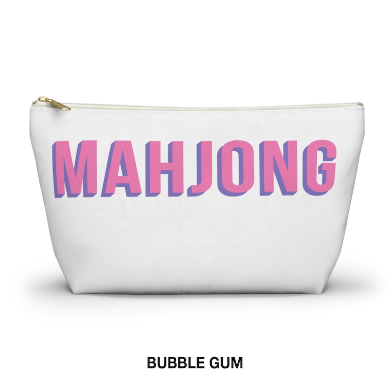 May include: A white zippered pouch featuring the word MAHJONG printed in large, blocky pink letters with a purple shadow effect. The accessory has a gold-toned metal zipper pull at the top left corner and a clean, minimalist design throughout.