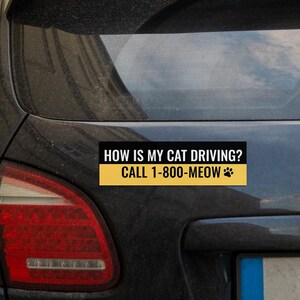 以下が含まれることがあります： 「HOW IS MY CAT DRIVING? CALL 1-800-MEOW」の白文字と足跡アイコンが入った黒いバンパーステッカー。テキストは黒い背景に黄色のストライプが入っています。