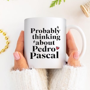 Puede incluir: Taza de cer&aacute;mica blanca con el texto "Probably thinking about Pedro Pascal" en negro. La taza tiene un asa blanca y la sostiene una persona que lleva un su&eacute;ter blanco. En el fondo se ve un collar de cuentas doradas.