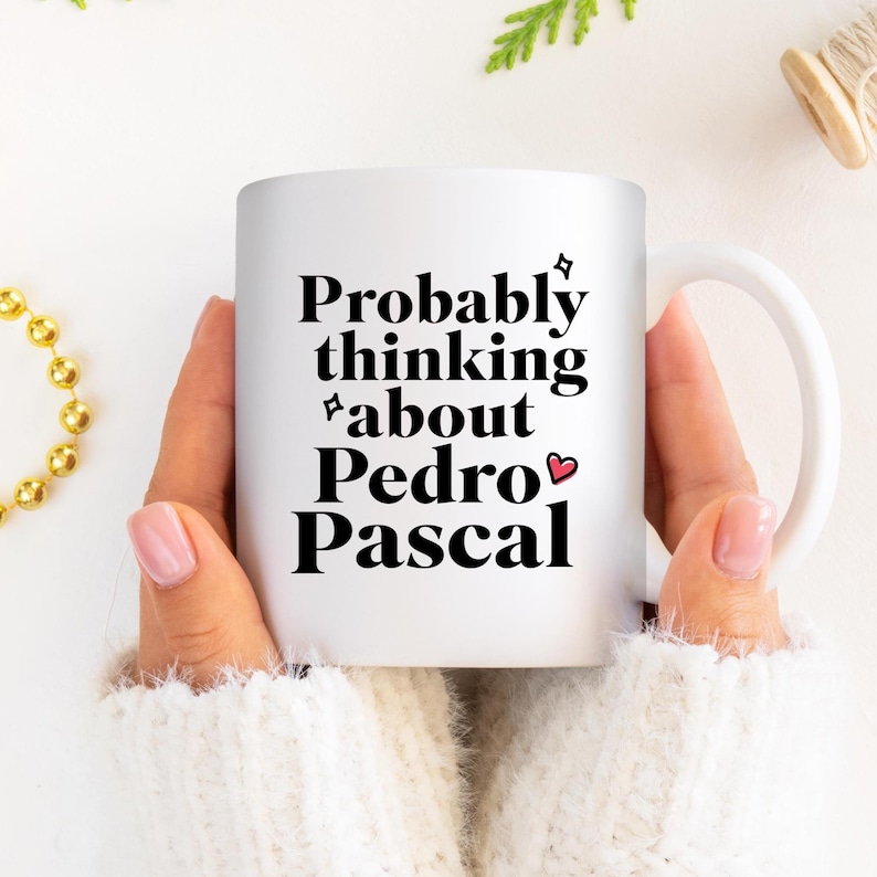 Puede incluir: Taza de cer&aacute;mica blanca con el texto "Probably thinking about Pedro Pascal" en negro. La taza tiene un asa blanca y la sostiene una persona que lleva un su&eacute;ter blanco. En el fondo se ve un collar de cuentas doradas.