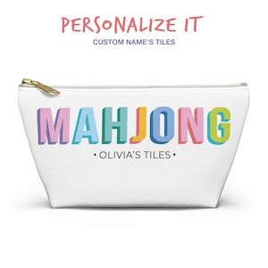 May include: A white zippered pouch featuring the word MAHJONG in large, colorful, three-dimensional block letters. Below the word, the text reads OLIVIA'S TILES in a simple black font. The pouch has a gold-toned metal zipper pull at the top.
