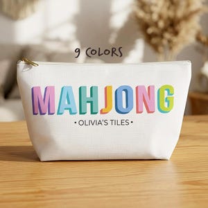 Può includere: Una pochette bianca con cerniera che presenta la parola MAHJONG in grandi lettere a blocchi 3D multicolori. Sotto la parola, il testo recita OLIVIA'S TILES in un semplice carattere nero. La pochette ha un cursore della cerniera dorato.