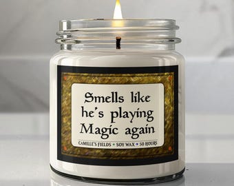 May include: A white soy wax candle in a clear glass jar with a lit wick. The label features a gold textured border and black text that reads: Smells like he's playing Magic again, Camille's Fields, Soy Wax, 50 Hours. The candle emits a warm glow.