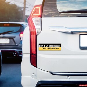 以下が含まれることがあります： 白い車のリアに貼られたステッカー。ステッカーには「HOW IS MY CAT DRIVING? CALL 1-800-MEOW」というテキストが黒で黄色い背景に書かれており、足跡が付いています。背景には他の車が見えます。