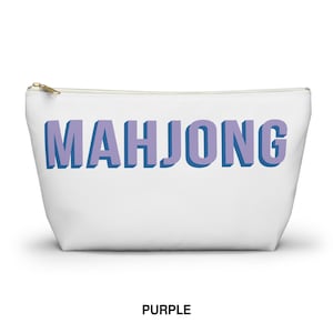 May include: A white zippered pouch featuring the word MAHJONG printed in large, block-style letters. The text has a light purple face with a dark blue shadow effect, creating a three-dimensional appearance against the plain white background.