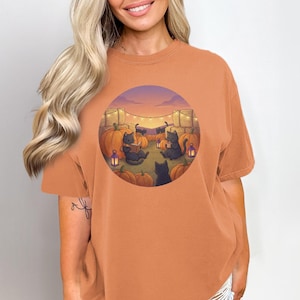 Op de afbeelding: Een roestoranje t-shirt met korte mouwen en een ronde grafische print. De illustratie toont verschillende zwarte katten die tussen oranje pompoenen en hooibalen zitten onder een lichtsnoer tijdens een zonsondergang.
