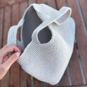 May include: A light gray, crocheted tote bag with two handles. The bag has a rounded shape and a textured surface. The interior of the bag is a darker gray color. The bag is shown on a wooden surface.