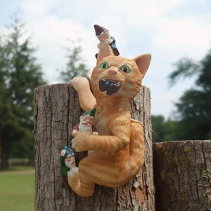 May include: A novelty garden ornament of an orange cat climbing a tree trunk, with three small gnome figures clinging to the cat. The cat has green eyes and an open mouth. The gnomes wear blue, green, and red outfits.