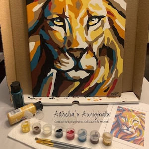 May include: A paint-by-numbers kit featuring a colorful abstract lion with a yellow, orange, and brown mane. The kit includes a canvas, paint tubes, brushes, and a reference image.