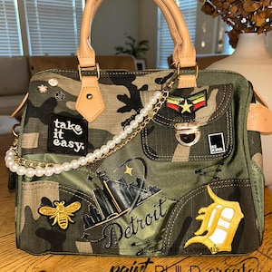 May include: Camouflage-print handbag with tan leather handles and details. Decorated with patches, pins, and a pearl chain. Features the text "take it easy," "Detroit," and "BE KIND." Includes a gold bee and a Detroit Tigers logo.