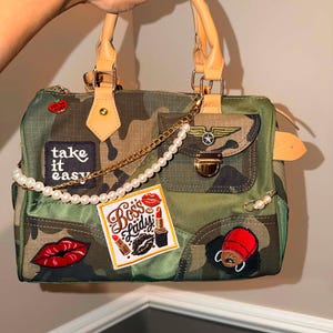 May include: A camouflage print handbag with tan handles and a gold chain accented with white pearls. The bag features various patches including text that reads take it easy and Boss Lady, along with images of red lips, a lipstick, and a cap.