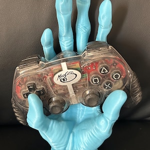 Könnte beinhalten: Eine hellblaue, im 3D-Druck hergestellte Hand hält einen transparenten schwarzen MadCatz-Videospielcontroller. Der Controller hat schwarze Knöpfe und ein silbernes MadCatz-Logo.