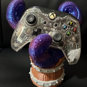May include: A clear Xbox controller with purple and blue accents, resting on a decorative barrel-shaped stand. The stand is brown with white spikes around the top and bottom. The controller has colored buttons and a black background.