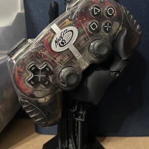 May include: A clear plastic video game controller with black buttons and a black and silver MadCatz logo. The controller is held by a black 3D printed hand.
