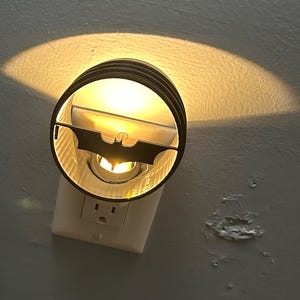Peut inclure: Un veilleuse en forme de silhouette de Batman noir et blanc branchée sur une prise électrique. La lumière est allumée et projette une ombre sur le mur.