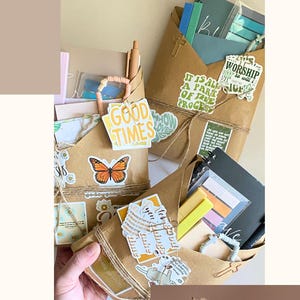 May include: A collection of brown paper envelopes filled with stationery, journals, and stickers featuring text like "GOOD TIMES" and "IT IS ALL A PART OF THE PROCESS." A Bible page with a gold cross paperclip and a metal charm is also visible.