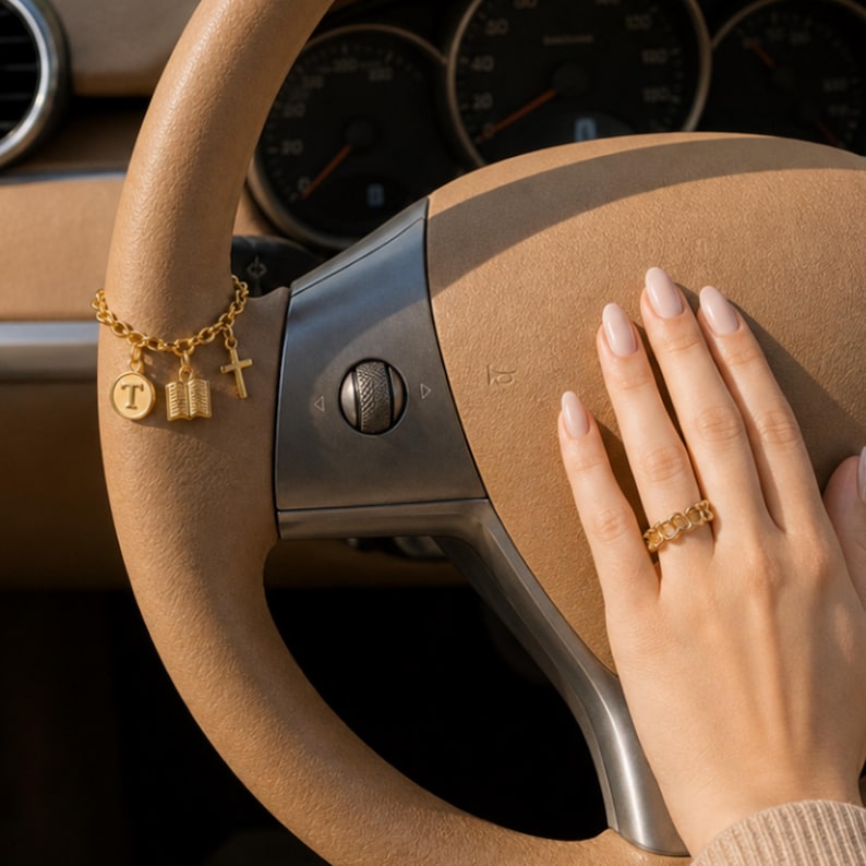 May include: A gold-toned charm bracelet featuring a circular pendant with the letter T, a small book charm, and a cross charm, alongside a gold-toned chain-link ring. The jewelry is displayed on a tan steering wheel with a metallic center hub.