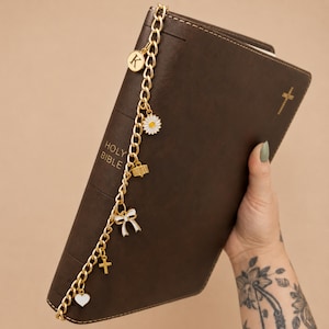 May include: A dark brown Holy Bible with a gold-toned metal chain bookmark attached to the spine. The chain features several charms including a letter K disc, a white daisy, a small book, a white bow, a cross, and a white heart charm.