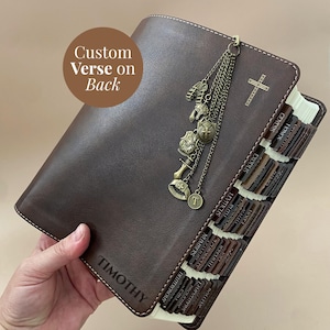 May include: A dark brown leather Bible cover featuring the name TIMOTHY embossed on the front, a gold cross emblem, and a bronze chain charm with armour-themed pendants. The cover includes dark brown index tabs for various books of the Bible.