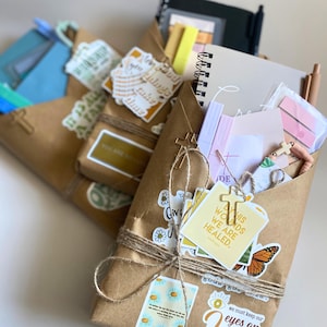 May include: Three brown paper packages tied with twine, each containing stationery items like notebooks, pens, and sticky notes. The packages are decorated with various stickers featuring floral designs, butterflies, and religious text including By His Wounds We Are Healed and God's Got This, along with small gold-colored cross charms.