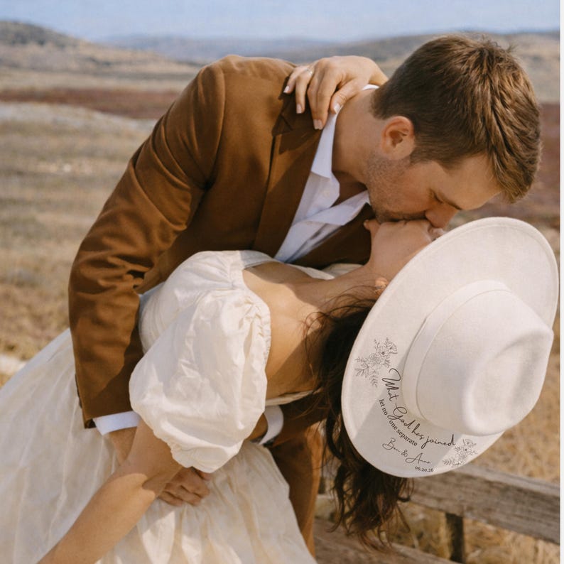 Puede incluir: Una pareja se abraza, la mujer lleva un sombrero blanco con detalles florales y texto. El hombre lleva una chaqueta marr&oacute;n y una camisa blanca. El sombrero dice "What God has joined, let no one separate, B & A, 06.04.23".