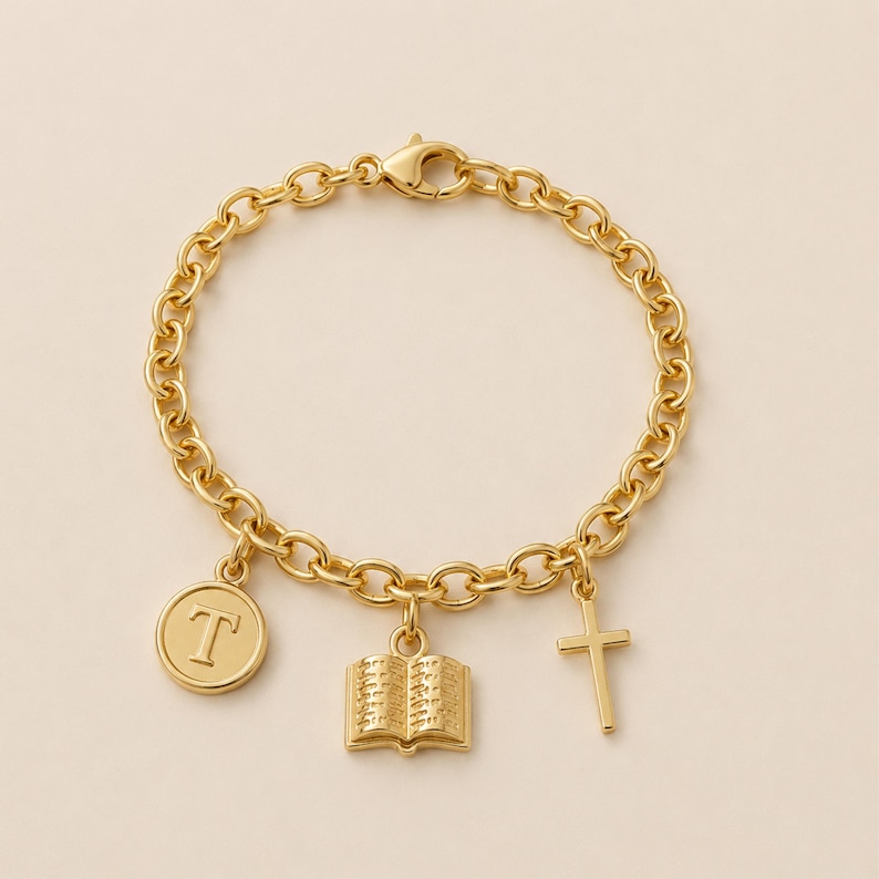 May include: A gold-tone chain link bracelet featuring three dangling charms: a circular disc with the letter T, an open book, and a simple cross. The bracelet is secured with a lobster claw clasp, showcasing a polished metallic finish throughout.