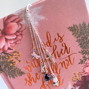 May include: A silver-toned metal bookmark featuring a curved top hook and four delicate hanging chains. The charms include a pink dragonfly, a silver cross, the letter K, a black cat with a red heart, and a pink tulip flower charm.