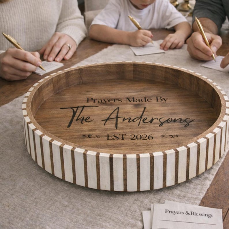 May include: A round wooden tray with a light brown interior and a white and brown striped exterior. The tray has the text "Prayers Made By The Andersons EST 2026" engraved on the inside. People are writing on paper with pens.