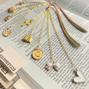May include: A gold-toned metal bookmark featuring a curved flat blade and multiple dangling gold chains. The chains are adorned with various charms including a cross, an open book, a daisy, a circular pendant with the letter C, a bow, and a heart.